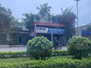 Bức ảnh chụp một con đường tại Việt Nam, có các cửa hàng kinh doanh san sát nhau. Dựa vào thông tin trên biển hiệu, địa điểm này là ở Định Bình, Yên Định, Thanh Hóa. Các biển hiệu có thể đọc được bao gồm: "CHĂN - GA - GỐI - ĐỆM SƠN NGUYỆT", "NHÀ SÁCH SƠN NGUYỆT" chuyên bán "SÁCH GIÁO KHOA - VĂN PHÒNG PHẨM", và các thương hiệu "THIÊN LONG" với khẩu hiệu "SỨC MẠNH TRI THỨC", "ANVICO" với khẩu hiệu "ẤM ÁP NHƯ LÒNG MẸ", và "NỆM KIM CƯƠNG". Hoạt động trong ảnh là cảnh giao thông thường ngày với một người đang đội mũ bảo hiểm và lái xe máy (xe máy) trên đường. Nhiều xe máy khác đang đỗ ven đường. Khung cảnh còn có cây cối, bụi rậm xanh tươi ở dải phân cách, một giàn hoa giấy màu hồng tím nổi bật và một cột đèn đường năng lượng mặt trời.。（日本語訳）。ベトナムのある通りを撮影した写真で、商店が軒を連ねています。看板の情報によると、この場所はタインホア省イエンディン県ディンビンです。読み取れる看板には、「SON NGUYET 掛け布団・シーツ・枕・マットレス」、「教科書・文房具」を専門に販売する「SON NGUYET書店」、そしてスローガン「知識の力」を掲げる「THIEN LONG」ブランド、「母の心のように温かい」を掲げる「ANVICO」ブランド、「KIM CUONGマットレス」などがあります。写真の中の活動は日常の交通風景で、一人の人物がヘルメットをかぶり、バイク（xe máy）を運転しています。他にも多くのバイクが道端に駐車されています。