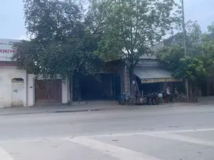 Bức ảnh chụp cảnh một con đường với các cửa hàng và nhà dân. Nổi bật là một cửa hàng kinh doanh xe đạp và xe điện có tên 'Thọ Nguyệt'. Phía trước cửa hàng, nhiều xe đạp và xe máy điện được xếp thành hàng. Một người đàn ông mặc áo ba lỗ màu trắng đang đứng gần đó. Có nhiều cây xanh lớn tỏa bóng mát xuống vỉa hè và một phần con đường nhựa. Trên các tòa nhà có một số biển hiệu với nội dung có thể đọc được như sau:。- Biển hiệu lớn bên trái, bị cây che một phần: 'THIẾT BỊ ĐIỆN TỬ - ĐIỆN KHUYẾ...', địa chỉ 'ĐC: CÂY XĂNG ĐỊNH...', và số điện thoại 'ĐT: 0982...'.。- Biển hiệu trên cửa hàng xe đạp: '...XE ĐẠP - ĐIỆN - MÁY...', 'BUÔN, BÁN LẺ XE ĐẠP - ĐIỆN...', '...G CHÍNH HIỆU'.。- Biển hiệu dọc trên cột nhà: 'CHUYÊN MUA BÁN TRAO ĐỔI XE ĐẠP XE ĐIỆN'.。- Biển hiệu màu vàng của cửa hàng: 'Thọ Nguyệt', 'ĐT: 01252 403548'.。- Trên tường có gắn biển số nhà '417'.。（日本語訳）。店舗や民家が立ち並ぶ通りの風景を写した写真です。目立っているのは「Thọ Nguyệt」という名前の自転車・電動バイク店です。店の前には、多くの自転車や電動バイクが並べられています。白いタンクトップを着た男性がその近くに立っています。大きな緑の木々が歩道とアスファルトの道路の一部に涼しい木陰を落としています。建物には、以下のように読み取れるいくつかの看板があります。- 左側の大きな看板、一部が木で隠れている：「THIẾT BỊ ĐIỆN TỬ - ĐIỆN KHUYẾ...」（電子機器 - 電...）、住所「ĐC: CÂY XĂNG ĐỊNH...」（住所：ディン...ガソリンスタンド）、電話番号「ĐT: 0982...」（電話：0982...）。- 自転車店の上の看板：「...XE ĐẠP - ĐIỆN - MÁY...」（...自転車 - 電動 - 機械...）、「BUÔN, BÁN LẺ XE ĐẠP - ĐIỆN...」（自転車・電動...の卸売、小売）、「...G CHÍNH HIỆU」（...正規品）。- 建物の柱にある縦長の看板：「CHUYÊN MUA BÁN TRAO ĐỔI XE ĐẠP XE ĐIỆN」（自転車・電動バイクの売買・交換専門）。- 店の黄色い看板：「Thọ Nguyệt」、「ĐT: 01252 403548」（電話：01252 403548）。- 壁には「417」という番地プレートが取り付けられています。