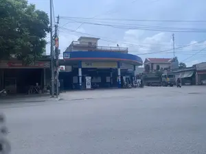 Bối cảnh là một con đường với một trạm xăng Petrolimex ở trung tâm. Trạm xăng có mái che màu xanh và cam. Trên mái che có các biển hiệu 'PETROLIMEX' và một biển hiệu lớn màu vàng ghi 'SH BẢO HIỂM ÔTÔ - XE MÁY', 'Bảo hiểm Sài Gòn - Hà Nội' cùng số điện thoại '1900 96 96 09'. Một bảng giá điện tử hiển thị các loại xăng như 'XĂNG RON 95-III' và 'XĂNG E5RON92-II'. Một vài người đang dừng xe máy tại các cột bơm xăng.。Bên trái trạm xăng là một cửa hàng, có thể là quán ăn, với biển hiệu màu đỏ ghi 'BÚN HUẾ CỐ ĐÔ'. Bên phải có một chiếc xe tải ben lớn đang đỗ. Khung cảnh xung quanh bao gồm các ngôi nhà, cây xanh, và nhiều cột điện với dây điện chằng chịt. Trên một cột điện có treo một lá cờ Phật giáo. Bầu trời có mây.。（日本語訳）。背景は、中央にペトロリメックスのガソリンスタンドがある道です。ガソリンスタンドには青とオレンジ色の屋根があります。屋根の上には「PETROLIMEX」の看板と、「SH BẢO HIỂM ÔTÔ - XE MÁY」、「Bảo hiểm Sài Gòn - Hà Nội」および電話番号「1900 96 96 09」と書かれた大きな黄色い看板があります。電子価格表示板には、「XĂNG RON 95-III」や「XĂNG E5RON92-II」といったガソリンの種類が表示されています。数人が給油機でバイクを停めています。ガソリンスタンドの左側には、おそらく飲食店と思われる店があり、「BÚN HUẾ CỐ ĐÔ」と書かれた赤い看板があります。右側には、一台の大きなダンプトラックが駐車しています。周囲の風景には、家々、緑の木々、そして電線が複雑に絡み合った多くの電柱が含まれています。一本の電柱には仏旗が掲げられています。空は曇っています。