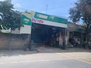 Nội dung văn bản trên các biển hiệu:。- Biển hiệu chính lớn: 'Castrol TƯ YẾN XE MÁY', bên dưới có dòng chữ 'SỬA CHỮA BẢO DƯỠNG THAY THẾ PHỤ TÙNG'.。- Biển hiệu dọc bên trái: 'Castrol TƯ YẾN XE MÁY', 'SỬA CHỮA, BẢO DƯỠNG', 'THAY THẾ PHỤ TÙNG'.。- Biển hiệu nhỏ bên trong cửa hàng: 'TƯ SỬA CHỮA BẢO DƯỠNG XE GA - XE [SỐ]', 'MUA BÁN XE'.。- Một phần biển hiệu 'PETROLIMEX' có thể nhìn thấy ở cửa hàng bên cạnh.。Mô tả bối cảnh và hoạt động:。Đây là hình ảnh chụp một tiệm sửa xe máy tên là 'Tư Yến' nằm ở ven đường. Cửa hàng có mặt tiền mở, bên trong chứa nhiều phụ tùng xe máy như lốp xe. Dựa trên thông tin từ các biển hiệu, cửa hàng này chuyên về sửa chữa, bảo dưỡng, thay thế phụ tùng và mua bán các loại xe máy. Cửa hàng cũng là đại lý hoặc sử dụng sản phẩm của thương hiệu dầu nhớt 'Castrol'.。Phía trước tiệm có một chậu cây cảnh. Con đường nhựa ở phía trước có vạch kẻ sọc chéo màu vàng. Cảnh vật xung quanh có cây xanh và bầu trời trong xanh. Trên mái nhà có một ăng-ten TV.。（日本語訳）。看板の文面内容：。- 大きなメイン看板：「Castrol トゥー・イェン バイク」、その下に「修理・メンテナンス・部品交換」の文字。- 左側の縦看板：「Castrol トゥー・イェン バイク」、「修理、メンテナンス」、「部品交換」。- 店内の小さな看板：「トゥー 修理・メンテナンス スクーター - マニュアル車」、「バイク売買」。背景と活動の説明：。これは道端にある「トゥー・イェン」という名前のバイク修理店を撮影した写真です。店は店先が開いており、中にはタイヤなどの多くのバイク部品が置かれています。看板の情報に基づくと、この店は各種バイクの修理、メンテナンス、部品交換、そして売買を専門としています。この店はまた、エンジンオイルブランド「カストロール」の代理店であるか、その製品を使用しています。店の前には観葉植物の鉢植えが一つあります。前のアスファルト道路には黄色の斜線が引かれています。周囲の景色には緑の木々と澄んだ青空があります。屋根の上にはテレビアンテナが一つあります。