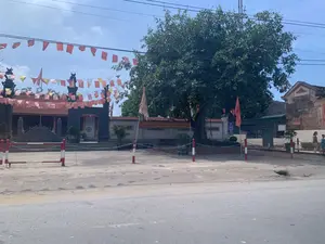 文字記録：壁には大きな赤い横断幕があり、白い文字で「革命の伝統を発揮し、幹部と人民は…新農村基準を満たす農村を建設する」と書かれている。右側の古い建物の壁には、「LUẬT SƯ 24H」という広告看板があり、電話番号は「1900.6574」である。より小さな看板には「Giữ Xe」という文字がある。風景と活動の描写：写真は、古い建築物の三関門の正面の光景を写したもので、村の亭（ディン）または歴史的遺跡である可能性がある。正面入口は、赤と白の縞模様に塗られた杭と鎖で一時的に封鎖されている。境内には大きな古木が一本立っている。右側では、一人の女性と一人の子供が、古い二階建ての家の隣の歩道を歩いている。前方の道はかなり閑散としている