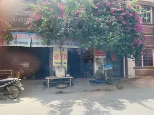 Hình ảnh chụp mặt tiền một cửa hàng ven đường, có thể ở một khu dân cư hoặc vùng nông thôn. Phía trước cửa hàng có một giàn hoa giấy lớn đang nở hoa màu hồng. Đây là một cơ sở kinh doanh đa dịch vụ, được thể hiện qua các biển hiệu: Biển hiệu lớn màu xanh lam ghi 'CHUYÊN: SỬA CHỮA MÁY BƠM NƯỚC - QUẠT ĐIỆN - NỒI CƠM ĐIỆN - DỤNG CỤ ĐIỆN CẦM TAY (KHOAN...)'. Một biển hiệu dọc màu vàng ghi 'HÀNG TẠP HÓA' và 'THUỐC LÀO'. Ngoài ra còn có biển hiệu 'ĐIỂM BÁN VIETTEL' và 'vinaphone'. Phía trước cửa hàng có một chiếc ghế tre, trên đó đặt một bao tải lớn màu trắng. Dưới đất có một cái rổ. Bên trái hình ảnh có một chiếc xe máy đang đỗ. Khung cảnh tĩnh lặng, không có hoạt động rõ rệt của con người.。（日本語訳）。道端にある店の正面を写した写真で、住宅地か田舎にあるのかもしれない。店の前には、ピンク色の花を咲かせている大きなブーゲンビリアの棚がある。これは多角的なサービスを提供する事業所であり、それは看板に示されている。青い大きな看板には「専門：ウォーターポンプ・扇風機・炊飯器・携帯電動工具（ドリルなど）の修理」と書かれている。黄色い縦長の看板には「雑貨」と「トゥオックラオ（ベトナムの刻みタバコ）」と書かれている。その他にも、「VIETTEL販売店」と「vinaphone」の看板がある。地面には籠が一つある。写真の左側にはバイクが一台停まっている。静かな光景で、はっきりとした人の活動は見られない。