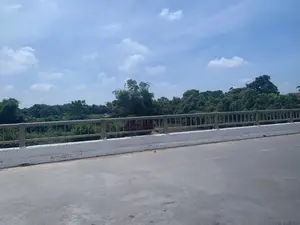 Bức ảnh chụp khung cảnh nhìn từ trên một cây cầu. Ở phía trước là mặt đường nhựa và lan can cầu được làm bằng bê tông sơn trắng, phía trên có tay vịn kim loại. Nhìn qua lan can là một khung cảnh làng quê với rất nhiều cây cối xanh tươi, rậm rạp. Có thể thấy một phần của một con sông hoặc kênh rạch chảy bên dưới cầu. Bầu trời trong xanh và có mây trắng. Không có người, phương tiện hay hoạt động cụ thể nào diễn ra. Không có bất kỳ văn bản hay biển báo nào trong ảnh.。（日本語訳）。写真は、ある橋の上から見た風景を撮影したものです。手前にはアスファルトの路面と、白いペンキで塗られたコンクリート製の橋の欄干があり、その上には金属製の手すりがあります。欄干の向こうには、青々とした木々が生い茂る田園風景が広がっています。橋の下を流れる川または運河の一部が見えます。空は青く澄んでおり、白い雲があります。人や乗り物、特定の活動は見られません。写真にはいかなる文字や標識もありません。