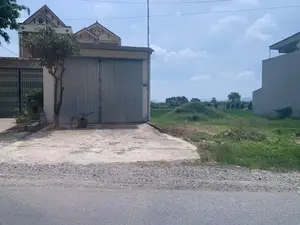 Bức ảnh chụp một khung cảnh ven đường ở một vùng nông thôn vào ban ngày. Bên trái là một ngôi nhà có sân bê tông và một cửa cuốn lớn bằng tôn. Trên tường cạnh cửa có gắn biển số nhà màu xanh ghi số '140', chữ bên dưới khó đọc. Phía trước nhà có trồng một vài cây xanh và chậu cây cảnh. Bên phải ngôi nhà là một cánh đồng lúa xanh mướt, ở giữa có một gò đất nhỏ. Nhìn ra xa là cây cối, một vài công trình kiến trúc khác và dãy núi dưới bầu trời xanh có mây. Con đường ở phía trước là đường nhựa. Khung cảnh chung rất yên tĩnh, không có người hay hoạt động nào đang diễn ra.。（日本語訳）。写真は、昼間の田舎の道端の風景を写しています。左側には、コンクリートの庭と大きなトタン製のシャッターがある家があります。ドアの横の壁には、「140」と書かれた青い番地プレートが取り付けられていますが、その下の文字は読みにくいです。家の前には、いくつかの緑の木と鉢植えの観葉植物が植えられています。家の右側には、青々とした水田が広がっており、その真ん中には小さな土の盛り上がりがあります。遠くには、木々、いくつかの他の建築物、そして雲のある青空の下に山脈が見えます。手前の道はアスファルトの道路です。全体的な風景はとても静かで、人や活動は見られません。