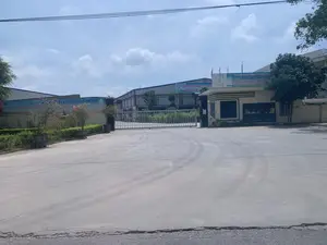 Đây là hình ảnh chụp lối vào của một nhà máy, có thể là một công ty may mặc, vào một ngày nắng. Cảnh vật bao gồm một cổng sắt lớn màu trắng đang đóng, một chốt bảo vệ ở bên phải và các tòa nhà nhà xưởng ở phía sau. Trên các tòa nhà và chốt bảo vệ có treo các băng rôn. Băng rôn bên phải trên chốt bảo vệ có nội dung: 'CÁN BỘ CNLĐ CÔNG TY TNHH MAY', khẩu hiệu '“ĐOÀN KẾT - TỰ TIN - SÁNG TẠO”', và 'XÂY DỰNG CÔNG TY PHÁT TRIỂN VÌ LỢI ÍCH NGƯỜI LAO ĐỘNG VÀ...'. Logo trên băng rôn có thể đọc là 'CÔNG TY TNHH MAY PHÚC SƠN'. Băng rôn bên trái ghi: 'CÁN BỘ, CÔNG NHÂN VIÊN CÔNG TY LÀM VIỆC BẰNG T...'. Một băng rôn khác ở xa hơn bên trái có chữ 'Kính Chào'. Xung quanh lối vào có cây xanh và các chậu cây cảnh. Sân trước cổng được trải bê tông rộng rãi, có vệt bánh xe trên mặt sân. Bầu trời trong xanh và có mây.。（日本語訳）。これは、ある晴れた日に撮影された、ある工場（おそらく縫製会社）の入り口の写真です。風景には、閉まっている大きな白い鉄の門、右側にある警備員詰所、そして後方にある工場の建物が含まれています。建物と警備員詰所には横断幕が掲げられています。右側の警備員詰所の上にある横断幕には、「縫製有限会社の幹部・労働者」、スローガン「“団結 - 自信 - 創造”」、そして「労働者の利益と…のために発展する会社を建設する」という内容が書かれています。横断幕のロゴは「PHUC SON縫製有限会社」と読めます。左側の横断幕には、「会社の幹部、従業員はT…で働く」と書かれています。さらに左奥にある別の横断幕には「歓迎」という文字があります。入り口の周りには緑の木々や鉢植えの観葉植物があります。門の前の庭は広々としてコンクリートで舗装されており、地面にはタイヤの跡があります。空は青く晴れ渡り、雲があります。