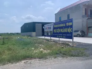 Bức ảnh chụp một khung cảnh ven đường, có thể ở khu vực ngoại ô hoặc nông thôn. Trung tâm của bức ảnh là một nhà kho lớn có vách và mái bằng tôn màu xanh lá cây. Cửa nhà kho đang mở. Bên cạnh nhà kho là một ngôi nhà hai tầng chưa hoàn thiện với tường bê tông. Phía trước có một tấm biển quảng cáo lớn màu xanh lam và trắng. Trên biển có ghi các thông tin sau: 'TỔNG KHO VĂN HOÀNG GIA - YÊN ĐỊNH', 'PHỤ KIỆN NGÀNH MỘC', và 'THIẾT BỊ NHÀ BẾP CAO CẤP BOSS', 'BỀN VỮNG THEO THỜI GIAN'. Biển cũng cung cấp địa chỉ 'THÔN ẤP TRÚ - XÃ ĐỊNH BÌNH - YÊN ĐỊNH - THANH HÓA', số điện thoại '082 965 1234', và website 'www.bossvn.vn'. Một chiếc ô tô SUV màu be đang đỗ gần ngôi nhà. Khu vực xung quanh là đất trống với cỏ dại và sỏi đá. Phía xa là cánh đồng và cây cối dưới bầu trời có mây.。（日本語訳）。写真は道端の風景を写したもので、郊外または田舎の地域である可能性があります。写真の中央には、緑色のトタンの壁と屋根を持つ大きな倉庫があります。倉庫の扉は開いています。倉庫の隣には、コンクリートの壁を持つ未完成の2階建ての家があります。前面には、青と白の大きな広告看板があります。看板には次の情報が書かれています：「TỔNG KHO VĂN HOÀNG GIA - YÊN ĐỊNH」（ヴァン・ホアン・ザー総合倉庫 - イエンディン）、「PHỤ KIỆN NGÀNH MỘC」（木工業用アクセサリー）、そして「THIẾT BỊ NHÀ BẾP CAO CẤP BOSS」（BOSS高級キッチン設備）、「BỀN VỮNG THEO THỜI GIAN」（時を超えて持続する）。看板には住所「THÔN ẤP TRÚ - XÃ ĐỊNH BÌNH - YÊN ĐỊNH - THANH HÓA」（タインホア省イエンディン県ディンビン社アプチュー村）、電話番号「082 965 1234」、ウェブサイト「www.bossvn.vn」も記載されています。ベージュ色のSUV車が家の近くに駐車しています。周囲は雑草や砂利のある空き地です。遠くには、曇り空の下に田んぼと木々が見えます。