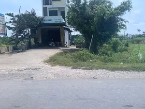 看板のテキスト内容：。左側の看板の内容：「Tuyết」、「シート張り替え」、「バイク修理」、「各種シート/カバー」、「洗車」、「電話番号：0349 411 426」。店舗の軒先の看板には、「バイク専門」、「自動車 - バイク」、「オイル交換」と書かれています。風景と活動：。写真は、ある農村地域の道端にあるバイク修理店を撮影したものです。店にはコンクリートの前庭があり、バイク修理、自動車とバイクのオイル交換、洗車、シート張り替えなどのサービスを提供しています。修理エリア内にバイクが1台停められています。店の隣には、畝に沿って青野菜が植えられた畑と多くの木々があります。後ろには近代的な多層階の家があります。遠くには木々、電柱、そして一つの建築物が見えます