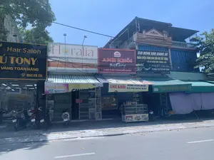 Bức ảnh chụp một dãy nhà phố thương mại liền kề trên một con đường ở thành phố Thanh Hóa vào một ngày nắng. Các cửa hàng có mặt tiền hướng ra đường, phía trên là các tầng nhà ở. Nhiều xe máy đang được đỗ trên vỉa hè. Hệ thống dây điện và cáp viễn thông chằng chịt phía trên các tòa nhà. Các biển hiệu có thể đọc được bao gồm:。- "Hair Salon Ú TONY", "VÀNG TOÀN NĂNG", SĐT: 0945 918 234.。- "Floralia Flower Boutique", chuyên thiết kế hoa sinh nhật, sự kiện, trang web www.floraliashop.com, SĐT: 0898.63.38.38.。- "Anh Khôi Bakery", với khẩu hiệu "LUÔN TƯƠI NGON MỖI NGÀY", địa chỉ "ĐC: 472 NGUYỄN TRÃI - P. PHÚ SƠN - TP. THANH HÓA". Phía trước tiệm có một tủ kính trưng bày bánh. Mái hiên có biển "ANH KHÔI BÁNH SINH NHẬT BÁNH KEM BẮP".。- "CLB BÓNG BÀN GIANG SƠ", chuyên "HUẤN LUYỆN BÓNG BÀN CHO MỌI ĐỐI TƯỢNG TỪ CƠ BẢN ĐẾN NÂNG CAO" và "TRAO ĐỔI - MUA BÁN DỤNG CỤ BÓNG BÀN", cũng tại địa chỉ "472 Nguyễn Trãi - P. Phú Sơn".。- Một biển hiệu nhỏ khác ghi "THẠNH HƯƠNG 472 Nguyễn Trãi".。- Một tấm biển đỏ "CHO THUÊ NHÀ" với số điện thoại liên hệ "LH: 0916 125 797" được treo trên một cửa sắt kéo đang đóng.。（日本語訳）。晴れた日にタインホア市のある通りで撮影された、隣接する商店街の家並みの写真。各店舗は通りに面しており、上階は住居となっている。多くのバイクが歩道に駐車されている。建物の上方には、電線や通信ケーブルが複雑に張り巡らされている。読み取れる看板には以下のものが含まれる：。- 「Hair Salon Ú TONY」、「VÀNG TOÀN NĂNG」、電話番号：0945 918 234。- 「Floralia Flower Boutique」、誕生日、イベント用の花のデザインを専門とし、ウェブサイトはwww.floraliashop.com、電話番号は0898.63.38.38。- 「Anh Khôi Bakery」、スローガンは「毎日いつでも新鮮で美味しい」、住所は「タインホア市フーソン区グエンチャイ通り472番地」。店の前にはケーキを陳列するガラスケースがある。軒先には「ANH KHÔI 誕生日ケーキ コーンクリームケーキ」という看板がある。- 「CLB BÓNG BÀN GIANG SƠ」（GIANG SƠ卓球クラブ）、「初心者から上級者まで、あらゆる対象者への卓球指導」および「卓球用具の交換・売買」を専門とし、こちらも住所は「グエンチャイ通り472番地 - フーソン区」。- 別の小さな看板には「THẠNH HƯƠNG グエンチャイ通り472番地」と書かれている。- 連絡先電話番号「LH: 0916 125 797」が記載された「CHO THUÊ NHÀ」（貸家）と書かれた赤い看板が、閉まっている鉄製のシャッターに掛けられている。