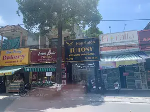 Bối cảnh là một con phố kinh doanh với nhiều cửa hàng nằm san sát nhau ở thành phố Thanh Hóa, dưới trời nắng. Trên vỉa hè có một cây xanh lớn, phía trước các cửa hàng có nhiều xe máy đang đậu. Một chiếc xe ba gác được dùng làm quầy hàng rong, chở đầy dưa hấu và dưa lưới để bán, trên xe có các biển ghi giá viết tay như "13 K 1kg" và "DƯA SIÊU NGỌT".。Các thông tin từ các biển hiệu có thể đọc được:。- "Bảo Trang Bakery" tại 476 Nguyễn Trãi, P. Phú Sơn, TP. Thanh Hóa, bán "BÁNH SINH NHẬT Á - ÂU", với biển "Giá Chỉ Từ 10k".。- "Sâm Bảo TRISO" tại 474 Nguyễn Trãi, với khẩu hiệu "DÂNG VUA - TIẾN CHÚA".。- "Hair Salon TÚ TONY" tại số 472B, một tiệm làm tóc với biển "KÉO VÀNG TOÀN NĂNG".。- "Floralia Flower Boutique", một cửa hàng hoa chuyên thiết kế hoa sinh nhật, chúc mừng.。- Cửa hàng "Thanh Hương" tại 472 Nguyễn Trãi.。- Một cửa hàng có biển hiệu màu đỏ ghi địa chỉ "ĐC: 472 NGUYỄN TRÃI".。- Một tấm biển "CHO THUÊ NHÀ" với số điện thoại liên hệ "LH: 0946 125 797" được treo trên cửa xếp.。（日本語訳）。背景は、晴天の下、タインホア市の商店街で、多くの店が隣接して並んでいます。歩道には大きな緑の木があり、店の前には多くのバイクが停まっています。一台の三輪車が移動販売の屋台として使われ、スイカとメロンをたくさん積んで販売しており、車には「1kg 13K」や「超甘いメロン」といった手書きの価格表示があります。読み取れる看板からの情報は以下の通りです：。- タインホア市フーソン区グエンチャイ通り476番地にある「Bảo Trang Bakery」は、「アジア・ヨーロッパ風バースデーケーキ」を販売しており、「価格はわずか10kから」という看板があります。- グエンチャイ通り474番地にある「Sâm Bảo TRISO」は、「王に捧げ、主に献上する」というスローガンを掲げています。- 472B番地にある「Hair Salon TÚ TONY」は、「万能の金のハサミ」という看板を掲げた美容室です。- 「Floralia Flower Boutique」は、誕生日やお祝いの花を専門にデザインする花屋です。- グエンチャイ通り472番地にある「Thanh Hương」店。- 「住所：グエンチャイ472番地」と記された赤い看板のある店。- シャッターに「貸家」という看板が掲げられており、連絡先電話番号「連絡先：0946 125 797」が記載されています。