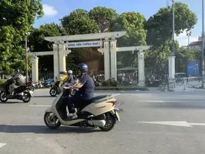 Bức ảnh chụp cảnh giao thông trên một con đường nhựa trước cổng vào một công viên. Trên cổng chính có dòng chữ 'CÔNG VIÊN THỐNG NHẤT'. Phía bên phải có một biển báo màu xanh nhưng chữ viết không rõ. Hoạt động chính là mọi người đang điều khiển xe máy tham gia giao thông, tất cả đều đội mũ bảo hiểm. Ở tiền cảnh, một người phụ nữ đang lái một chiếc xe tay ga màu be. Phía sau cổng công viên là không gian có nhiều cây xanh lớn, và có một vài người đang đi bộ và ngồi nghỉ gần lối vào.。（日本語訳）。写真は、ある公園の入り口ゲート前のアスファルト道路上の交通風景を撮影したものです。正門には「CÔNG VIÊN THỐNG NHẤT」という文字があります。右側には青い看板がありますが、文字ははっきり見えません。主な活動は、人々がバイクを運転して交通に参加していることで、全員がヘルメットを着用しています。前景には、一人の女性がベージュ色のスクーターを運転しています。公園のゲートの後ろには、大きな緑の木々がたくさんある空間が広がっており、入り口の近くで数人が歩いたり座って休んだりしています。