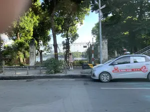 Bức ảnh chụp một khung cảnh đường phố ven hồ. Ở phía trước, một chiếc ô tô màu bạc đang di chuyển trên đường nhựa. Thân xe có dán decal màu đỏ với các dòng chữ và số, có thể nhận ra một phần số điện thoại '3.888.123' và số '23'. Bên cạnh đường là vỉa hè, nơi một người phụ nữ mặc trang phục màu hồng đang đi bộ. Vỉa hè được bao bọc bởi hàng rào sắt và các cột trụ bằng đá. Phía sau hàng rào là một không gian có nhiều cây xanh và một hồ nước lớn. Xa xa phía bên kia hồ có thể thấy lờ mờ vài tòa nhà cao tầng. Bầu trời trong xanh và có nắng, cho thấy đây là một ngày thời tiết đẹp. Có một cột sơn sọc đỏ trắng, có thể là cọc tiêu giao thông, đứng trên vỉa hè.。（日本語訳）。写真は湖畔の街の風景を写しています。手前では、銀色の自動車がアスファルトの道路を走行しています。車体には文字と数字が書かれた赤いデカールが貼られており、電話番号の一部「3.888.123」と数字の「23」が認識できます。道路の隣には歩道があり、そこではピンク色の服を着た女性が歩いています。歩道は鉄柵と石の柱で囲まれています。柵の後ろには緑豊かな木々と大きな湖がある空間が広がっています。湖の向こう側遠くに、いくつかの高層ビルがぼんやりと見えます。空は青く晴れており、天気の良い日であることがわかります。歩道には、交通用の標柱と思われる、赤と白の縞模様に塗られた柱が立っています。