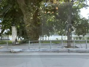 Bức ảnh được chụp từ một phương tiện đang di chuyển, nhìn ra một con đường ven hồ. Khung cảnh là một công viên hoặc khu vực công cộng với nhiều cây xanh lớn. Có một vỉa hè lát gạch và một lan can kim loại thấp chạy dọc theo nó. Phía sau lan can là một hàng rào kim loại cao hơn, ngăn cách với khu vực công viên. Trong công viên, có những cây cổ thụ, một trong số đó được gia cố bằng khung kim loại. Trên thân cây này có vẻ như có sơn số màu vàng nhưng không thể đọc rõ. Một cột trụ bằng đá có chữ viết nhưng đã bị mờ và không đọc được. Ở phía xa, qua hàng cây là mặt hồ nước. Có một vài người đang đi bộ và tập thể dục trên con đường ven hồ.。（日本語訳）。風景は、大きな緑の木々がたくさんある公園または公共エリアです。舗装された歩道と、それに沿って低い金属製の手すりがあります。手すりの後ろには、公園エリアとを隔てる、より高い金属製のフェンスがあります。公園内には古い木があり、そのうちの1本は金属製のフレームで補強されています。文字が書かれた石の柱がありますが、かすれていて読めません。遠く、木々の向こうには湖面が見えます。湖畔の道では、数人が散歩や運動をしています。