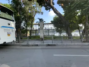 Bức ảnh chụp một khung cảnh ven đường vào một ngày nắng. Bên trái ảnh là phần đuôi của một chiếc xe khách lớn màu trắng. Cạnh đó là vỉa hè và một hàng rào sắt chạy dài, ngăn cách với một khu vực có vẻ là công viên ven hồ. Trên một trụ cột bê tông của hàng rào có các chữ và hình vẽ graffiti, trong đó có thể đọc được chữ "VAXE". Phía sau hàng rào là một hồ nước rộng và có nhiều cây xanh cổ thụ. Có thể thấy một vài người đang đi dạo trên con đường ven hồ. Bầu trời trong xanh, cho thấy thời tiết đẹp.。（日本語訳）。写真は、晴れた日の道端の風景を写しています。写真の左側には、白い大型バスの後部があります。その隣には歩道と、湖畔の公園と思われるエリアを隔てる長く続く鉄柵があります。柵のコンクリートの柱の一つにはグラフィティの文字と絵があり、その中には「VAXE」という文字が読み取れます。柵の後ろには広い湖と、緑豊かな古木がたくさんあります。湖畔の道で散歩している数人の人々が見えます。空は青く澄んでおり、天気が良いことを示しています。