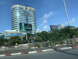 Bối cảnh là một khu vực đô thị hiện đại dưới bầu trời xanh quang đãng. Đối tượng chính trong ảnh là một tòa nhà văn phòng cao tầng có thiết kế hình trụ độc đáo, với bề mặt chủ yếu là kính. Trên tòa nhà này có gắn nhiều biển hiệu và logo của các công ty. Trên đỉnh là logo 'VIMC' và dòng chữ 'TỔNG CÔNG TY HÀNG HẢI VIỆT NAM'. Các tầng dưới có biển hiệu của 'vuanem', 'SIEMENS', 'DAIKIN', 'XE MIỀN BẮC HOÀNG DŨNG', và 'GMO-Z.com RUNSYSTEM'.。Ở khối nhà thấp hơn liền kề, có biển hiệu 'ACET TRUNG TÂM ĐÀO TẠO ANH NGỮ ÚC'. Một biển quảng cáo lớn được dựng lên, quảng bá cho 'BIA SAIGON SPECIAL' với dòng chữ 'VỊ SPECIAL CHO DỊP SPECIAL'. Phía sau, một tòa nhà khác có logo 'FWD' cũng có thể được nhìn thấy.。Cảnh quan đường phố bao gồm một con đường nhựa, dải phân cách hoặc lề đường có lan can kim loại, nhiều cây xanh, và các chậu cây cảnh. Vỉa hè được sơn hai màu đỏ và trắng. Bức ảnh ghi lại một khung cảnh tĩnh của thành phố, không có hoạt động cụ thể nào của con người đang diễn ra.。（日本語訳）。背景は、澄み切った青空の下の近代的な都市部です。写真の主な被写体は、ユニークな円筒形のデザインをした高層オフィスビルで、表面は主にガラス張りです。このビルには、多くの企業の看板やロゴが取り付けられています。最上部には「VIMC」のロゴと「TỔNG CÔNG TY HÀNG HẢI VIỆT NAM」の文字があります。下の階には、「vuanem」、「SIEMENS」、「DAIKIN」、「XE MIỀN BẮC HOÀNG DŨNG」、「GMO-Z.com RUNSYSTEM」の看板があります。隣接する低い方の建物には、「ACET TRUNG TÂM ĐÀO TẠO ANH NGỮ ÚC」の看板があります。大きな広告看板が設置されており、「VỊ SPECIAL CHO DỊP SPECIAL」という文言で「BIA SAIGON SPECIAL」を宣伝しています。後方には、「FWD」のロゴがある別の建物も見えます。街の風景には、アスファルトの道路、金属製の手すりのある中央分離帯または路肩、多くの緑樹、観賞用の植木鉢が含まれています。歩道は赤と白の二色で塗られています。この写真は、特定の人間活動が見られない、静かな都市の光景を捉えています。