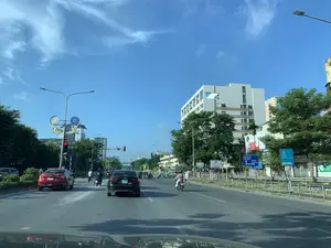Một con đường đô thị lớn, nhiều làn xe vào ban ngày dưới bầu trời xanh trong. Hai bên đường là cây xanh và các tòa nhà cao tầng. Hoạt động chính là giao thông với nhiều ô tô và xe máy đang di chuyển hoặc dừng chờ đèn đỏ (đèn tín hiệu đang hiển thị số 42). Một chiếc ô tô màu đỏ có dán chữ 'TẬP LÁI' và 'flydoimoi.com'. Các biển báo chỉ đường màu xanh lam chỉ hướng đến một bến xe, một đường vành đai, và một bệnh viện cách đó 100m. Ngoài ra còn có các biển hiệu và biển quảng cáo với nội dung như 'Vietcombank', 'Sinocare', 'SÁCH CHUYÊN CHỮA...', 'Bao Cham', 'GỘI ĐẦU DƯỠNG SINH', và 'CHĂM SÓC DA'. Trên cột đèn tín hiệu giao thông có một cụm trang trí nghệ thuật hình con thuyền với họa tiết tròn.。（日本語訳）。澄んだ青空の下、日中の多くの車線がある大きな都市の道路。道路の両側には緑の木々と高層ビルがある。主な活動は交通で、多くの自動車やバイクが移動しているか、あるいは赤信号で停止して待っている（信号機は数字の42を表示している）。一台の赤い自動車には「TẬP LÁI」と「flydoimoi.com」という文字が貼られている。青い道路標識が、バスターミナル、環状道路、そして100m先にある病院への方向を示している。その他にも、「Vietcombank」、「Sinocare」、「SÁCH CHUYÊN CHỮA...」、「Bao Cham」、「GỘI ĐẦU DƯỠNG SINH」、「CHĂM SÓC DA」といった内容の看板や広告看板がある。信号機の柱の上には、丸い模様のある船の形をした芸術的な装飾群がある。