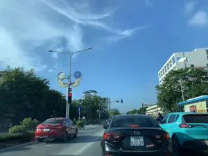 Bối cảnh là một con đường nhiều làn xe trong một thành phố, được chụp từ bên trong một chiếc ô tô. Các phương tiện giao thông đang dừng lại chờ đèn đỏ dưới trời nắng đẹp, bầu trời trong xanh có vài gợn mây.。Ở phía trước, có một chiếc ô tô màu đen hiệu Mazda 3, trên xe có dán chữ 'SKYACTIV' và một nhãn dán ghi '© DÂN CHỦ'.。Bên trái là một chiếc ô tô màu đỏ, phía sau xe có dán biển 'TẬP LÁI' và trang web 'Taylaimoi.com'.。Bên phải là một chiếc ô tô màu xanh ngọc và một phần của một chiếc xe buýt. Trên thân xe buýt có quảng cáo lớn cho sản phẩm 'SÂM NHUNG' và dòng chữ 'CÔNG TY CỔ PHẦN DƯỢC PHẨM TRUNG ƯƠNG 3'.。Cột đèn giao thông đang báo hiệu đèn đỏ, với đồng hồ đếm ngược kỹ thuật số hiển thị số '14'. Cột đèn này được trang trí bằng các họa tiết hình chiếc thuyền cách điệu và các hình tròn, gợi liên tưởng đến hoa văn trên trống đồng.。Hai bên đường là hàng cây xanh và các tòa nhà hiện đại.。（日本語訳）。背景は、ある都市の複数車線のある道路で、車内から撮影されたものです。車両は美しい晴天の下、赤信号で停止しており、空は青く澄み、いくつかの雲が浮かんでいます。前方には、黒いマツダ3が1台あり、車には「SKYACTIV」の文字と「© DÂN CHỦ」と書かれたステッカーが貼られています。左側には赤い車が1台あり、車の後部には「TẬP LÁI」（練習中）の表示とウェブサイト「Taylaimoi.com」が貼られています。右側には、翡翠色の車とバスの一部が見えます。バスの車体には、「SÂM NHUNG」（高麗人参と鹿茸）という製品の大きな広告と、「CÔNG TY CỔ PHẦN DƯỢC PHẨM TRUNG ƯƠNG 3」（中央第三製薬株式会社）という文字が書かれています。信号機は赤信号で、デジタル式のカウントダウンタイマーが「14」という数字を表示しています。この信号機の柱は、様式化された船の形や円形の模様で装飾されており、銅鼓の文様を連想させます。道路の両側には、緑の並木と近代的な建物があります。