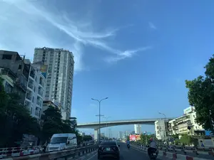 Bức ảnh ghi lại khung cảnh giao thông trên một con đường đô thị lớn ở Việt Nam vào một ngày đẹp trời, bầu trời trong xanh với vài gợn mây. Con đường có nhiều làn xe, dải phân cách cứng ở giữa, và đông đúc các phương tiện như ô tô và xe máy đang di chuyển. Một cây cầu vượt hiện đại bắc ngang qua con đường ở phía xa. Dọc hai bên đường là các tòa nhà cao tầng san sát. Về chữ viết, có một tòa nhà chung cư/văn phòng nổi bật ở bên trái với dòng chữ 'HOA PHAT' màu xanh trên nóc, và một tòa nhà khác ở phía xa có chữ 'VINACCO'. Hoạt động chính được ghi lại là dòng chảy giao thông đô thị với nhiều người đang điều khiển phương tiện.。（日本語訳）。この写真は、ベトナムのある大きな都市の道路の交通風景を、晴れた日に撮影したものです。空は青く澄み渡り、雲が少し浮かんでいます。道路には多くの車線があり、中央には頑丈な分離帯が設置され、自動車やバイクなどの車両がひしめき合って走行しています。遠くには、近代的な高架橋が道路を横切っています。道路の両脇には、高層ビルが立ち並んでいます。文字については、左側に目立つマンション/オフィスビルがあり、その屋上には青い文字で「HOA PHAT」と書かれています。また、遠くにある別のビルには「VINACCO」という文字が見えます。記録されている主な活動は、多くの人々が車両を運転している都市の交通の流れです。