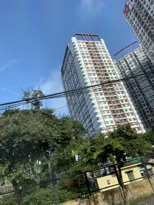 Bức ảnh chụp từ góc thấp hướng lên các tòa nhà chung cư cao tầng hiện đại dưới bầu trời trong xanh. Các tòa nhà có màu trắng chủ đạo với các chi tiết trang trí màu đỏ cam ở ban công và trên tầng thượng. Bầu trời xanh biếc, có một vài vệt mây trắng mỏng và một vệt dài có thể do máy bay tạo ra. Phía tiền cảnh có nhiều cây xanh, một số bị nhòe do chuyển động, cho thấy ảnh có thể được chụp từ một phương tiện đang di chuyển. Một mạng lưới dây điện dày đặc vắt ngang qua khung hình. Phía dưới có thể thấy một phần hàng rào và tường. Đây là một khung cảnh đô thị.。（日本語訳）。青空の下、近代的な高層マンションを見上げるようにローアングルから撮影された写真。建物は白を基調とし、バルコニーと最上階にオレンジがかった赤色の装飾が施されている。空は真っ青で、薄い白い雲がいくつかあり、飛行機によってできたと思われる長い筋が一本ある。前景には多くの緑の木々があり、その一部は動きによってぶれていることから、移動中の乗り物から撮影された可能性があることがわかる。密集した電線網がフレームを横切っている。これは都市の風景である。