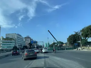 Bản ghi chữ viết:。- Biển hiệu trên tòa nhà màu vàng: "CÔNG TY CP NHỮNG TRANG VÀNG VIỆT NAM", "YELLOW PAGES JSC".。- Chữ trên cần cẩu: "GIA KHÁNH 0913 297 455".。- Biển chỉ đường màu xanh lam: "PHÒNG CÔNG CHỨNG", bên dưới có chữ "SỞ TƯ PHÁP TP. HÀ NỘI" và chỉ dẫn khoảng cách.。- Các biển hiệu quảng cáo khác có thể nhìn thấy: "Funiki", "TẬP ĐOÀN HÒA PHÁT", "incco".。Mô tả bối cảnh và hoạt động:。- Địa điểm: Khung cảnh trên một con đường lớn, nhiều làn xe tại một khu vực đô thị sầm uất ở Việt Nam. Bên phải là một công trường xây dựng lớn.。- Hoạt động: Các phương tiện bao gồm ô tô và xe máy đang tham gia giao thông đông đúc. Một chiếc cần cẩu lớn đang hoạt động tại công trường.。- Vật thể: Nhiều loại ô tô, xe máy, các tòa nhà cao tầng hai bên đường, cần cẩu, silo xi măng, hàng rào công trường, cột đèn giao thông và cây xanh. Bầu trời trong xanh với vài gợn mây.。（日本語訳）。文字の記録：。- 黄色い建物の看板：「CÔNG TY CP NHỮNG TRANG VÀNG VIỆT NAM」、「YELLOW PAGES JSC」。- クレーン上の文字：「GIA KHÁNH 0913 297 455」。- 青い案内標識：「PHÒNG CÔNG CHỨNG」、その下には「SỞ TƯ PHÁP TP. HÀ NỘI」の文字と距離の案内がある。背景と活動の説明：。- 場所：ベトナムの賑やかな都市部にある、複数車線の大通りの風景。右側には大規模な建設現場がある。- 活動：自動車やバイクなどの車両が混雑した交通に参加している。建設現場では大型クレーンが稼働している。- 物体：様々な種類の自動車、バイク、道路両側の高層ビル、クレーン、セメントサイロ、建設現場のフェンス、信号機の柱、そして緑の木々。空は青く澄んでおり、雲が少し浮かんでいる。