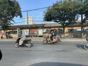 Bối cảnh là một con đường nhựa đông đúc tại một thành phố ở Việt Nam. Nhiều người đang tham gia giao thông bằng xe máy. Nổi bật có một cặp đôi đi trên chiếc xe máy số màu đỏ, chở theo nhiều túi hàng hóa gồm rau củ, cho thấy họ có thể vừa đi chợ về. Cả hai đều đội mũ bảo hiểm. Một người phụ nữ khác mặc bộ đồ chống nắng màu xám đang điều khiển một chiếc xe tay ga màu đỏ. Bên lề đường có một nhà chờ xe buýt, nơi có một người đàn ông đang đứng xem điện thoại. Trên nhà chờ có một biển quảng cáo lớn với dòng chữ 'Dầu nhớt số 1 của người Việt' và thương hiệu 'AT POWER'. Phía sau nhà chờ là một hàng rào, bên trong có vẻ là một bến xe hoặc bãi đỗ xe với nhiều xe khách. Ở phía xa, có thể nhìn thấy các tòa nhà chung cư cao tầng dưới bầu trời xanh.。（日本語訳）。背景はベトナムのある都市の、交通量の多いアスファルトの道路です。多くの人々がバイクで交通に参加しています。目立っているのは、赤いカブ型バイクに乗ったカップルで、野菜を含む多くの品物が入った袋を運んでおり、市場から帰ってきたところかもしれません。二人ともヘルメットをかぶっています。別の女性が灰色のUVカットウェアを着て、赤いスクーターを運転しています。道端にはバスの待合所があり、そこでは一人の男性が立って携帯電話を見ています。待合所の上には、「ベトナム人のためのNo.1エンジンオイル」という文言と「AT POWER」というブランド名が書かれた大きな広告看板があります。待合所の後ろにはフェンスがあり、その内側は多くの長距離バスがあるバスターミナルか駐車場のようです。遠くには、青空の下に高層マンションが見えます。
