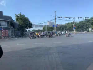 Bối cảnh là một ngã tư đường phố đông đúc vào ban ngày, dưới bầu trời trong xanh. Rất nhiều người đang điều khiển xe máy và một vài chiếc ô tô đang dừng lại, có vẻ như đang chờ đèn tín hiệu giao thông. Hầu hết người đi xe máy đều đội mũ bảo hiểm. Có một người chở một chiếc thùng hàng màu vàng ở yên sau xe máy. Hai bên đường có cây xanh và các tòa nhà. Ở phía xa có thể thấy một tòa nhà chung cư cao tầng. Hệ thống dây điện chằng chịt trên các cột điện. Bên lề đường bên trái, có hai tấm biển báo tuyên truyền an toàn giao thông màu đỏ với nội dung: 'KHÔNG SỬ DỤNG ĐIỆN THOẠI KHI ĐANG ĐIỀU KHIỂN PHƯƠNG TIỆN GIAO THÔNG' và 'TUÂN THỦ ĐÚNG TỐC ĐỘ QUY ĐỊNH KHI LÁI XE'. Ngoài ra còn có các biển báo giao thông khác như biển báo cấm đi ngược chiều và biển chỉ dẫn làn đường.。（日本語訳）。背景は、青空の下、日中の混雑した交差点です。多くの人々がバイクを運転しており、数台の自動車が停止しています。交通信号を待っているようです。バイクに乗っている人のほとんどはヘルメットを着用しています。バイクの後部座席に黄色い荷物箱を載せている人が一人います。道路の両側には緑の木々と建物があります。遠くには高層の集合住宅が見えます。電柱の上では電線が複雑に絡み合っています。左側の路肩には、交通安全を啓発する2つの赤い標識があり、内容は「車両運転中の携帯電話使用禁止」と「運転時は規定速度を遵守せよ」です。その他にも、逆走禁止の標識や車線案内標識など、他の交通標識もあります。