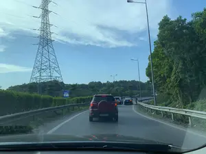 Bức ảnh được chụp từ bên trong một chiếc ô tô đang di chuyển trên một con đường có nhiều làn xe, có thể là quốc lộ hoặc đường cao tốc. Phía trước có một hàng dài các xe ô tô, trong đó chiếc gần nhất là một chiếc SUV màu đỏ. Con đường được bao bọc bởi các dải hộ lan tôn sóng bằng kim loại. Bên trái đường có một cột điện cao thế bằng kết cấu thép rất lớn. Hai bên đường là cây cối và bụi rậm xanh tươi. Bầu trời trong xanh với vài gợn mây. Ở phía xa bên trái có một biển báo giao thông màu xanh với vạch chéo đỏ, đây là biển báo hiệu "Hết khu đông dân cư".。（日本語訳）。この写真は、複数の車線がある道路、おそらく国道か高速道路を走行中の車内から撮影されたものです。前方には長い車の列があり、その中で最も手前にあるのは赤いSUVです。道路は金属製の波形ガードレールで囲まれています。道路の左側には、非常に大きな鉄骨構造の高圧送電鉄塔があります。道路の両側には、青々とした木々や茂みがあります。空は青く澄んでおり、いくつかの筋雲があります。左手の遠くに、青地に赤い斜線の入った交通標識があります。これは「人口密集地域終わり」を示す標識です。