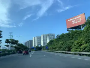 Bản ghi văn bản từ hình ảnh:。Trên biển quảng cáo lớn màu đỏ bên phải có dòng chữ: "MỜI QUẢNG CÁO" và hai số điện thoại "0965 77 8118", "0346 07 3287".。Các biển báo giao thông màu xanh chỉ dẫn hướng đi đến "CẦU GIẼ", "CẦU THANH TRÌ", "CẦU THĂNG LONG". Một biển báo khác chỉ "LỐI VÀO 300m" đi "PHÁP VÂN - CẦU GIẼ". Phía xa có một biển quảng cáo mang tên "XINGFA".。Mô tả cảnh quan và hoạt động:。Bối cảnh là một con đường cao tốc hoặc đường vành đai hiện đại với nhiều làn xe. Một số ô tô đang di chuyển trên đường. Dọc hai bên đường là hàng cây xanh và lan can bảo vệ. Ở phía sau, có thể thấy một dãy các tòa nhà chung cư cao tầng, cho thấy đây là một khu vực đô thị phát triển. Bên trái có một cột điện cao thế. Bầu trời trong xanh, quang đãng. Hoạt động chính là giao thông đường bộ.。（日本語訳）。画像からのテキスト記録：。右側にある大きな赤い広告看板には、「広告募集」という文字と、「0965 77 8118」、「0346 07 3287」という2つの電話番号が書かれています。青い交通標識が「ザウゼー橋」、「タインチー橋」、「タンロン橋」への方向を示しています。別の標識は、「ファップヴァン - ザウゼー」方面へ行く「入口 300m」を示しています。遠くには「XINGFA」という名前の広告看板があります。風景と活動の描写：。背景は、多くの車線を持つ現代的な高速道路または環状道路です。何台かの車が道路を走行しています。道路の両側には、緑の並木とガードレールがあります。後方には、高層マンション群が見え、ここが発展した都市部であることを示しています。左側には高圧電線の鉄塔があります。空は青く、澄み渡っています。主な活動は道路交通です。