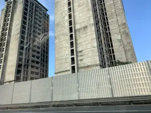 Bức ảnh chụp hai tòa nhà cao tầng bằng bê tông cốt thép, trông có vẻ như bị bỏ hoang hoặc đang xây dựng dở dang. Các tòa nhà không có cửa sổ hay lớp sơn hoàn thiện, để lộ ra kết cấu bê tông thô và các ô cửa trống. Ở phía trước là một hàng rào chắn bằng kim loại, có thể là hàng rào cách âm của một con đường cao tốc, cho thấy bức ảnh được chụp từ một phương tiện đang di chuyển. Bầu trời trong xanh và không có hoạt động nào của con người được nhìn thấy. Không có bất kỳ chữ viết hay biển hiệu nào trong ảnh.。（日本語訳）。鉄筋コンクリート製の2つの高層ビルが写っている写真で、廃墟か建設途中のように見えます。建物には窓や仕上げの塗装がなく、むき出しのコンクリート構造と空の窓枠が露出しています。手前には金属製の防護柵があり、高速道路の遮音壁である可能性があり、写真が移動中の乗り物から撮影されたことを示唆しています。空は青く澄んでおり、人の活動は見られません。写真にはいかなる文字や看板もありません。