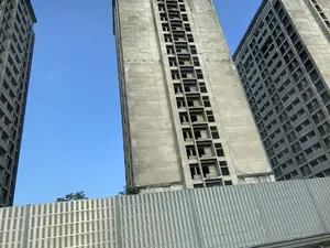 Bức ảnh chụp một góc nhìn từ dưới lên các tòa nhà cao tầng bằng bê tông. Tòa nhà ở trung tâm và các tòa nhà xung quanh dường như đang trong quá trình xây dựng dở dang hoặc bị bỏ hoang. Phần kết cấu thô đã hoàn thành nhưng chưa được lắp đặt cửa sổ hay hoàn thiện mặt ngoài, để lộ ra nhiều ô trống đen. Bề mặt các tòa nhà là bê tông trần, trông cũ kỹ. Ở phía trước, có một hàng rào chắn màu xám, có thể là một bức tường cách âm dọc theo một con đường lớn. Bầu trời trong xanh. Không có người hay hoạt động nào diễn ra. Không có bất kỳ văn bản hay biển báo nào có thể đọc được.。（日本語訳）。中央の建物と周囲の建物は、建設途中であるか、放棄されているように見えます。躯体構造は完成していますが、窓の設置や外壁の仕上げはまだ行われておらず、多くの黒い開口部が露出しています。建物の表面は打ちっ放しのコンクリートで、古びて見えます。手前には灰色のフェンスがあり、それは大きな道路に沿った防音壁かもしれません。空は青く澄んでいます。人や活動の様子は見られません。読み取れる文字や看板は一切ありません。