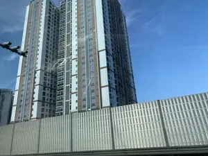Bức ảnh chụp một tòa nhà chung cư cao tầng, hiện đại dưới bầu trời trong xanh. Tòa nhà có kiến trúc thẳng đứng, sơn màu trắng kết hợp với các mảng màu nâu và xám, có nhiều cửa sổ và ban công. Ở phía trước tòa nhà là một bức tường chống ồn lớn bằng kim loại, cho thấy địa điểm có thể nằm cạnh một con đường lớn hoặc đường cao tốc trên cao. Phía bên trái của bức ảnh có một cột gắn các camera giám sát. Trên một tòa nhà khác ở phía xa bên trái, có thể nhìn thấy một phần dòng chữ "ECOCI..." nhưng không rõ toàn bộ nội dung. Khung cảnh cho thấy đây là một khu đô thị phát triển, và bức ảnh dường như được chụp từ bên trong một phương tiện đang di chuyển.。（日本語訳）。青空の下、近代的で高層の集合住宅を撮影した写真です。建物は垂直的な建築様式で、白色を基調に茶色と灰色の部分が組み合わされており、多くの窓とバルコニーがあります。建物の手前には大きな金属製の防音壁があり、この場所が主要道路や高架高速道路の隣にある可能性を示しています。写真の左側には、監視カメラが取り付けられた柱があります。左奥の別の建物には、「ECOCI...」という文字の一部が見えますが、全文ははっきりしません。この光景は、ここが発展した都市部であることを示しており、写真は移動中の乗り物の中から撮影されたようです。