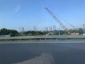 Bức ảnh được chụp từ một phương tiện đang di chuyển trên đường cao tốc, nhìn qua lan can đường là một công trường xây dựng. Trong công trường có một chiếc cần cẩu bánh xích lớn màu vàng và một máy xúc. Nhiều vật liệu xây dựng như dầm thép được xếp trên mặt đất. Ở phía sau là khu vực cây xanh, và xa hơn là đường chân trời của một khu đô thị với nhiều tòa nhà chung cư cao tầng và các cột điện cao thế. Bầu trời trong xanh, quang đãng. Toàn cảnh cho thấy một khu vực đang trong quá trình đô thị hóa và xây dựng.。（日本語訳）。現場には、一台の大きな黄色のクローラークレーンと一台のショベルカーがある。鉄骨などの多くの建設資材が地面に積まれている。空は青く、澄み渡っている。全景は、都市化と建設が進んでいる地域を示している。