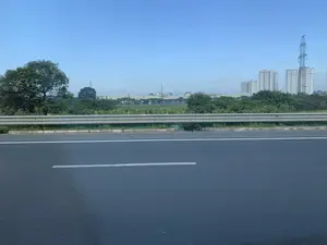 Bức ảnh chụp quang cảnh từ một con đường lớn hoặc đường cao tốc. Ở phía gần là mặt đường nhựa có vạch kẻ đường màu trắng và một dải hộ lan bằng kim loại. Phía bên kia đường là một dải cây xanh. Nhìn ra xa là một khung cảnh kết hợp giữa đô thị và công nghiệp, bao gồm nhiều tòa nhà chung cư cao tầng màu trắng, một cột điện cao thế lớn, và các công trình có mái bằng trông giống nhà xưởng. Bầu trời trong xanh, không có mây.。（日本語訳）。大きな道路または高速道路からの風景を撮影した写真です。手前には、白い車線が引かれたアスファルトの路面と、金属製のガードレールがあります。道路の向こう側には、緑の木々の帯があります。空は青く澄んでおり、雲はありません。