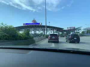 Bối cảnh là một trạm thu phí trên đường cao tốc, được chụp từ góc nhìn bên trong một chiếc ô tô. Phía trước có hai chiếc ô tô, một chiếc màu đỏ và một chiếc màu đen, đang di chuyển về phía các làn thu phí. Đây là trạm thu phí trên cao tốc Pháp Vân - Cầu Giẽ, được quản lý bởi 'CÔNG TY CỔ PHẦN BOT PHÁP VÂN - CẦU GIẼ' như được ghi trên biển hiệu điện tử lớn. Biển hiệu này cũng cung cấp thông tin đường dây nóng của Cảnh sát Giao thông: 'THÔNG TIN ĐƯỜNG DÂY NÓNG TRỰC BAN CSGT, SĐT: 02283899666 - ĐỘI 3 P6 CỤC CSGT'. Các biển báo treo phía trên các làn xe cho biết đây là 'LÀN THU PHÍ TỰ ĐỘNG KHÔNG DỪNG (ETC)'. Ở phía xa bên phải, có một tấm biển quảng cáo cho 'SƠN ĐẠI BÀNG'. Khung cảnh cho thấy hoạt động giao thông đang diễn ra bình thường trên đường cao tốc dưới bầu trời xanh, quang đãng.。（日本語訳）。背景は高速道路の料金所で、車内からの視点で撮影されています。前方には赤と黒の2台の車があり、料金レーンに向かって進んでいます。これはファップヴァン-カウゼー高速道路の料金所で、大きな電光掲示板に書かれているように「PHAP VAN - CAU GIE BOT株式会社」によって管理されています。この掲示板には交通警察のホットライン情報も記載されています：「交通警察当直ホットライン情報、電話番号：02283899666 - 交通警察局第6部第3隊」。車線の上に掲げられた標識は、ここが「ノンストップ自動料金収受レーン（ETC）」であることを示しています。遠く右側には、「SƠN ĐẠI BÀNG」の広告看板があります。この光景は、青く澄み渡った空の下、高速道路で交通が正常に行われていることを示しています。