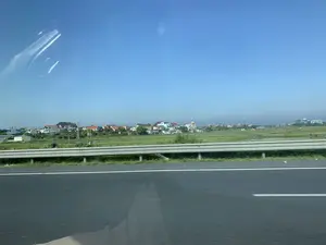 Bức ảnh ghi lại khung cảnh nông thôn Việt Nam, được chụp từ một phương tiện đang di chuyển trên đường cao tốc. Ở tiền cảnh là lan can đường bằng kim loại. Phía sau lan can là những cánh đồng xanh mướt, có thể là ruộng lúa, và một khu dân cư ở phía xa. Ngôi làng bao gồm nhiều ngôi nhà với mái ngói đỏ và cam, xen kẽ với các công trình hiện đại hơn. Có thể nhìn thấy một tháp chuông nhà thờ giữa các ngôi nhà. Bầu trời trong xanh, quang đãng. Không có chữ viết nào có thể đọc được trong ảnh.。（日本語訳）。この写真はベトナムの田園風景を記録したもので、高速道路を移動中の乗り物から撮影された。前景には金属製のガードレールがある。ガードレールの後ろには、おそらく水田であろう青々とした田畑と、遠くに住宅地が広がっている。その村は、赤やオレンジ色の瓦屋根の家々が多く、より近代的な建物も点在している。空は青く、澄み渡っている。写真には読み取れる文字はない。