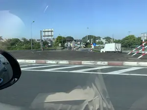 Bức ảnh được chụp từ bên trong một chiếc ô tô đang di chuyển trên đường cao tốc vào một ngày trời trong xanh. Phía trước là một trạm thu phí đường bộ với nhiều làn xe. Trên cổng trạm có các biển hiệu điện tử, có thể đọc được dòng chữ 'LÀN THU PHÍ ETC' và 'LÀN XỬ LÝ SỰ CỐ', cho thấy đây là trạm thu phí tự động không dừng. Có một số phương tiện giao thông khác như ô tô con và một chiếc xe tải thùng màu trắng đang tiến vào khu vực trạm thu phí. Dọc theo đường có các cột đèn, biển báo giao thông, và các cọc tiêu, rào chắn sọc đỏ trắng. Hai bên đường là cây cối và nhà cửa. Góc dưới bên trái của ảnh là một phần của gương chiếu hậu ô tô.。（日本語訳）。この写真は、晴れた日に高速道路を走行中の車内から撮影されたものです。前方には、複数の車線がある料金所があります。料金所のゲートの上には電光掲示板があり、「ETC料金レーン」と「トラブル処理レーン」という文字が読み取れ、これがノンストップ自動料金収受システムであることを示しています。乗用車や白い箱型トラックなど、他の車両も料金所エリアに進入しています。道路沿いには、街灯、交通標識、そして赤と白の縞模様のポールやガードフェンスがあります。道路の両側には木々や家々が見えます。