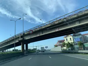 Bức ảnh được chụp từ góc nhìn của người ngồi trong xe ô tô đang di chuyển trên một đường cao tốc. Phía trước là một cây cầu vượt bằng bê tông bắc ngang qua đường. Trên dầm cầu có gắn một biển báo giao thông hình tròn, viền đỏ, nền trắng, ghi rõ giới hạn chiều cao là "4,3m". Dưới gầm cầu và trên các trụ đỡ có các vạch sơn phản quang màu đỏ-trắng và vàng-đen để cảnh báo. Bầu trời quang đãng, có màu xanh và mây trắng. Dọc theo đường cao tốc là hệ thống cột đèn chiếu sáng. Có nhiều phương tiện khác đang lưu thông trên đường, bao gồm xe tải và xe ô tô con. Bên phải đường là các tòa nhà và một tấm biển quảng cáo có chữ "MUNIC", các chữ khác không thể đọc rõ.。（日本語訳）。写真は、高速道路を走行中の車内にいる人の視点から撮影されたものです。前方には、道路を横切るコンクリート製の高架橋があります。橋桁には、赤枠に白地の円形交通標識が設置されており、「4.3m」という高さ制限が明記されています。橋の下と橋脚には、警告のために赤白および黄黒の反射塗装が施されています。空は晴れ渡り、青空に白い雲が浮かんでいます。高速道路沿いには、照明灯の支柱が並んでいます。道路上には、トラックや乗用車を含む他の多くの車両が通行しています。道路の右側には建物と、「MUNIC」という文字が書かれた看板があり、他の文字ははっきりと読み取れません。