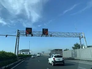 Bức ảnh ghi lại quang cảnh giao thông trên một tuyến đường cao tốc hiện đại ở Việt Nam vào một ngày nắng đẹp, trời trong xanh. Hình ảnh được chụp từ góc nhìn của người ngồi trong xe ô tô. Trên đường có nhiều phương tiện đang di chuyển, nổi bật là một chiếc xe VinFast Fadil màu trắng ở làn bên phải, phía trước có một xe khách và một xe tải. Con đường có nhiều làn được phân chia rõ ràng bằng vạch kẻ đường. Hai bên đường có tường chống ồn và dải phân cách trồng cây xanh. Phía trên, một giàn kết cấu thép lớn bắc ngang qua đường, có gắn hai bảng hiệu điện tử. Các bảng hiệu này đang hiển thị thông tin bằng đèn LED, tạo thành hình mặt cười màu đỏ, có thể là tín hiệu cho thấy giao thông đang thuận lợi. Các chữ viết trên bảng hiệu điện tử và các biển quảng cáo ở xa không thể đọc rõ.。（日本語訳）。写真は、晴れて青空が広がる美しい日に、ベトナムのある近代的な高速道路の交通風景を捉えたものである。この写真は、車内に座っている人の視点から撮影されたものである。道路上には多くの車両が走行しており、右車線には白いVinFast Fadilが目立っている。その前方にはバス1台とトラック1台がいる。道路は複数の車線があり、車線で明確に区切られている。道路の両側には防音壁があり、中央分離帯には緑が植えられている。これらの掲示板はLEDライトで情報を表示しており、赤いスマイリーフェイスの形を成している。これは交通が順調であることを示す信号かもしれない。電光掲示板や遠くの広告看板の文字は、はっきりと読み取ることができない。