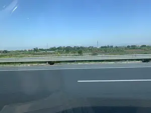 Bức ảnh được chụp từ một phương tiện đang di chuyển trên đường cao tốc, nhìn ra ngoài cửa sổ. Cảnh vật cho thấy một con đường nhựa với vạch kẻ đường màu trắng, được ngăn cách bởi một dải lan can hộ lan bằng kim loại. Phía sau lan can là một khung cảnh nông thôn đặc trưng với các thửa ruộng, vườn chuối và cây cối xanh tươi. Ở phía xa, có thể thấy những ngôi nhà dân và các cột điện cao thế dưới bầu trời trong xanh. Không có chữ viết hay hoạt động cụ thể nào của con người được ghi lại, chỉ có cảnh quan thiên nhiên và cơ sở hạ tầng.。（日本語訳）。写真は、高速道路を移動中の乗り物の窓から撮影されたものです。風景には、白い車線のあるアスファルト道路が写っており、金属製のガードレールで隔てられています。ガードレールの後ろには、田畑、バナナ園、そして青々とした木々がある、特徴的な田園風景が広がっています。遠くには、青空の下に民家や高圧電線の鉄塔が見えます。