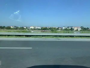 Bức ảnh được chụp từ một phương tiện đang di chuyển trên đường cao tốc, nhìn ra khung cảnh bên ngoài. Ở phía trước là mặt đường nhựa và một dải hộ lan bằng kim loại. Phía sau dải hộ lan là một khu dân cư ở vùng nông thôn hoặc ngoại ô, với nhiều ngôi nhà có kiến trúc đa dạng nằm xen kẽ giữa những hàng cây xanh. Có một cái ao hoặc hồ nước nằm giữa đường và khu dân cư. Bầu trời trong xanh, quang đãng. Có thể thấy hình ảnh phản chiếu của một phần nội thất xe ô tô trên cửa kính.。（日本語訳）。高速道路を走行中の乗り物から撮影された写真で、外の景色を眺めている。手前にはアスファルトの路面と金属製のガードレールがある。ガードレールの後ろには、田舎または郊外の住宅地があり、多様な建築様式の多くの家が緑の木々の間に点在している。道路と住宅地の間には、池か湖がある。空は青く澄み渡っている。