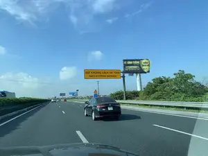 Bức ảnh chụp cảnh giao thông trên một tuyến đường cao tốc ở Việt Nam dưới bầu trời xanh và có mây. Nhiều ô tô đang di chuyển trên đường, trong đó có một chiếc xe màu đen ở làn bên phải. Dọc đường có các biển báo và biển quảng cáo. Một biển báo màu vàng bắc ngang đường ghi 'GIỮ KHOẢNG CÁCH AN TOÀN' và 'SAFE STOPPING DISTANCE'. Gần đó có biển báo 'CHIỀU CAO AN TOÀN 5m'. Bên phải có một biển quảng cáo lớn của 'VĨNH TƯỜNG SAINT-GOBAIN' cho sản phẩm 'TRẦN THẠCH CAO SIÊU THANH LỌC KHÍ' với các thông tin 'SIÊU BẢO VỆ', 'BẢO VỆ 5 LỚP' và số điện thoại '0984 99 11 22'. Ở phía xa hơn có thể thấy các biển quảng cáo của 'ỐNG THÉP HÒA PHÁT SỐ 1 VIỆT NAM' và 'THÉP VIỆT ĐỨC'. Hai bên đường có lan can bảo vệ và cây xanh.。（日本語訳）。青く雲のある空の下、ベトナムのある高速道路の交通風景を撮影した写真です。多くの自動車が道路を走行しており、その中には右車線に一台の黒い車がいます。道路沿いには標識や広告看板があります。道路を横切る黄色い標識には「GIỮ KHOẢNG CÁCH AN TOÀN」と「SAFE STOPPING DISTANCE」と書かれています。その近くには「CHIỀU CAO AN TOÀN 5m」という標識があります。右側には、「VĨNH TƯỜNG SAINT-GOBAIN」の製品「TRẦN THẠCH CAO SIÊU THANH LỌC KHÍ」の大きな広告看板があり、「SIÊU BẢO VỆ」、「BẢO VỆ 5 LỚP」、そして電話番号「0984 99 11 22」という情報が記載されています。さらに遠くには、「ỐNG THÉP HÒA PHÁT SỐ 1 VIỆT NAM」と「THÉP VIỆT ĐỨC」の広告看板が見えます。道路の両側にはガードレールと緑の木々があります。