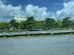 Bức ảnh được chụp từ một phương tiện đang di chuyển trên đường, nhìn ra một khu vực có vẻ là khu công nghiệp. Ở phía trước là mặt đường nhựa và một dải lan can kim loại, trên đó có dòng chữ phun sơn có thể đọc được một phần là "Lh 094475325...". Phía sau lan can là một hàng cây xanh. Ở hậu cảnh, có một tòa nhà nhà máy lớn với logo "CJ" và biểu tượng ba màu đặc trưng (xanh, đỏ, cam). Xung quanh tòa nhà có các cấu trúc công nghiệp, cột điện và đường dây điện. Bầu trời trong xanh và có nhiều mây trắng.。（日本語訳）。写真は、道路を移動中の乗り物から撮影されたもので、工業地帯と思われる区域を眺めています。手前にはアスファルトの路面と金属製のガードレールがあり、その上には「Lh 094475325...」と部分的に読み取れるスプレー塗装の文字があります。ガードレールの後ろには、緑の木々が並んでいます。背景には、「CJ」のロゴと特徴的な3色（青、赤、オレンジ）のシンボルがある大きな工場の建物があります。建物の周りには、工業用の建造物、電柱、そして電線があります。空は青く澄んでおり、白い雲が多くあります。