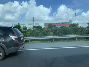 Bức ảnh được chụp từ một phương tiện đang di chuyển trên đường cao tốc. Phía bên trái là phần đuôi của một chiếc ô tô SUV màu đen đang chạy song song. Con đường được ngăn cách với khu vực bên cạnh bởi một dải hộ lan bằng kim loại. Phía sau dải hộ lan là một hàng cây xanh. Ở phía xa là một tòa nhà lớn, hiện đại, có vẻ là một nhà máy hoặc khu công nghiệp, với các bức tường màu trắng và đỏ. Bầu trời trong xanh với nhiều mây trắng. Có các cột điện và đường dây điện trong khung cảnh. Không có văn bản nào trên biển báo hoặc công trình có thể đọc được.。（日本語訳）。写真は高速道路を移動中の乗り物から撮影されたものです。左側には、並走している黒いSUV車の後部があります。道路は金属製のガードレールによって隣接するエリアと隔てられています。ガードレールの後ろには緑の木々が並んでいます。遠くには、白と赤の壁を持つ、工場か工業団地と思われる大きな近代的な建物があります。空は青く、白い雲がたくさん浮かんでいます。風景の中には電柱と電線があります。標識や建物には読み取れる文字はありません。