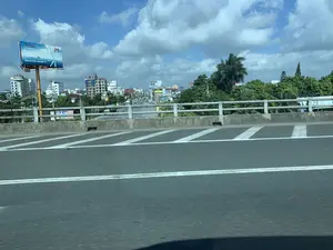 Bức ảnh ghi lại quang cảnh một thành phố ở Việt Nam, được chụp từ trên một cây cầu hoặc đường trên cao khi đang di chuyển. Ở phía trước là mặt đường nhựa với các vạch kẻ đường màu trắng và lan can cầu. Phía xa là khung cảnh đô thị với nhiều tòa nhà, cây xanh và các phương tiện giao thông đang di chuyển trên đường phố bên dưới.。Về chữ viết, có một biển quảng cáo lớn nổi bật ở bên trái của công ty PTSC (A member of PETROVIETNAM) với khẩu hiệu 'TRỌN GIẢI PHÁP, VẸN NIỀM TIN' và logo kỷ niệm '30 NĂM 1993-2023'. Dưới biển quảng cáo có ghi số điện thoại '0977 44 55 62'. Ngoài ra, có thể nhìn thấy các biển hiệu khác ở xa hơn như 'HOTEL TÙNG NAM' và một biển hiệu cho 'NHÀ NGHỈ'.。Cảnh vật bao gồm các tòa nhà cao tầng, trong đó có một công trình đang xây dựng với giàn giáo bên ngoài. Nhiều cây xanh, bao gồm cả cây cọ, mọc xen kẽ giữa các tòa nhà. Bầu trời trong xanh và có nhiều mây trắng. Hoạt động chính là giao thông đô thị với ô tô, xe tải và xe máy đang lưu thông.。（日本語訳）。写真はベトナムのある都市の光景を記録したもので、移動中の橋または高架道路の上から撮影されたものです。手前には、白い車線のあるアスファルトの路面と橋の欄干があります。遠方には、多くの建物、緑樹、そして下の通りを走行する交通車両からなる都市景観が広がっています。文字に関しては、左側にPTSC社（ペトロベトナムのメンバー）の大きな広告看板が目立っており、「完全なソリューション、揺るぎない信頼」というスローガンと、「30年 1993-2023」の記念ロゴがあります。広告看板の下には、電話番号「0977 44 55 62」が書かれています。さらに、遠くには「HOTEL TÙNG NAM」や「ゲストハウス」用の看板も見ることができます。景物には高層ビルが含まれており、その中には外側に足場が組まれた建設中の建物が一つあります。ヤシの木を含む多くの緑樹が、建物の間に点在して生えています。空は青く澄んでおり、白い雲が多く浮かんでいます。主な活動は都市交通であり、自動車、トラック、バイクが通行しています。