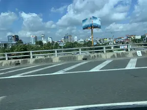 Bức ảnh chụp quang cảnh một khu vực đô thị từ trên một cây cầu hoặc đường cao tốc, nhìn từ một phương tiện đang di chuyển. Ở phía trước là mặt đường nhựa có vạch kẻ đường chéo màu trắng và một dải phân cách bằng bê tông. Phía sau lan can cầu là khung cảnh một thành phố với nhiều tòa nhà cao tầng, cây xanh và một tháp nước. Nổi bật trong số đó là một tòa nhà đang trong quá trình xây dựng, được bao bọc bởi giàn giáo. Bầu trời trong xanh, có nhiều mây trắng.。Các thông tin bằng chữ có thể đọc được bao gồm:。- Trên một biển quảng cáo lớn, hình hộp: logo "PTSC", dòng chữ kỷ niệm "30 NĂM 1993-2023" và khẩu hiệu "TRỌN GIẢI PHÁP, VỮNG NIỀM TIN".。- Trên một tòa nhà: "TỔNG CÔNG TY ĐẦU TƯ PHÁT TRIỂN ĐƯỜNG CAO TỐC VIỆT NAM".。- Các biển hiệu khác ở xa: "HOTEL HÙNG SƠN", "A.ĐÔNG".。- Một cột mốc bên đường ghi "H.8" và "278".。（日本語訳）。写真は、橋または高速道路の上から、移動中の乗り物から見た都市部の光景を撮影したものです。手前には、白い斜線の道路標示があるアスファルトの路面と、コンクリートの中央分離帯があります。橋の欄干の後ろには、多くの高層ビル、緑の木々、そして給水塔がある街の風景が広がっています。その中で目立っているのは、足場で覆われた建設中の建物です。空は青く澄んでおり、白い雲が多く浮かんでいます。読み取れる文字情報は以下の通りです：。- 大きな箱型の広告看板には、「PTSC」のロゴ、記念の文字「30 NĂM 1993-2023」、そしてスローガン「TRỌN GIẢI PHÁP, VỮNG NIỀM TIN」があります。- ある建物には、「TỔNG CÔNG TY ĐẦU TƯ PHÁT TRIỂN ĐƯỜNG CAO TỐC VIỆT NAM」と書かれています。- 遠くにある他の看板：「HOTEL HÙNG SƠN」、「A.ĐÔNG」。- 道路脇のキロポストには「H.8」と「278」と記されています。