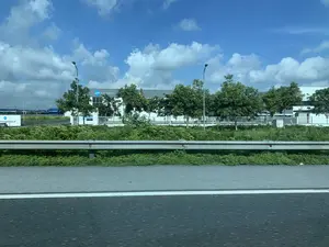 Bức ảnh chụp cảnh quan từ một phương tiện đang di chuyển trên đường cao tốc vào một ngày trời trong xanh, có mây trắng. Bên cạnh đường là một lan can bảo vệ bằng kim loại và một dải cây cỏ xanh. Ở phía xa là một tòa nhà nhà máy lớn, hiện đại, có tường màu trắng và xám. Có một biển hiệu màu trắng ở cổng ghi 'CÔNG TY TNHH SẢN PHẨM SHOFU VIỆT NAM'. Trên tòa nhà cũng có logo và dòng chữ 'SHOFU PRODUCTS VIETNAM'. Xung quanh nhà máy được bao bọc bởi hàng rào trắng và có trồng nhiều cây xanh. Một số cột đèn đường cũng xuất hiện trong khung cảnh.。（日本語訳）。白い雲のある晴れた日に、高速道路を移動中の乗り物から撮影された風景写真です。道路の脇には金属製のガードレールと緑の草地があります。遠くには、白と灰色の壁を持つ、大きくて近代的な工場ビルがあります。門には「CÔNG TY TNHH SẢN PHẨM SHOFU VIỆT NAM」と書かれた白い看板があります。ビルの上にも「SHOFU PRODUCTS VIETNAM」というロゴと文字があります。工場の周りは白いフェンスで囲まれ、多くの緑の木が植えられています。いくつかの街灯も風景の中に現れています。