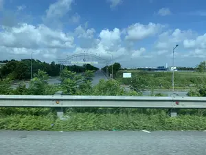 Bức ảnh chụp quang cảnh lối vào một khu công nghiệp, nhìn từ một phương tiện đang di chuyển. Trên một cổng chào lớn bằng kim loại bắc qua đường, có dòng chữ 'KHU CÔNG NGHIỆP ĐỒNG VĂN II MỞ RỘNG' và dòng chữ nhỏ hơn bên dưới 'SẴN SÀNG ĐỒNG HÀNH CÙNG CÁC NHÀ ĐẦU TƯ'. Đây là một con đường nhựa rộng, nhiều làn xe, hai bên trồng cây xanh và có hệ thống đèn đường. Bầu trời trong xanh và có nhiều mây. Ở phía xa có thể thấy các tòa nhà nhà xưởng. Bên phải đường có một biển báo giao thông giới hạn tốc độ 30 km/h. Một chiếc ô tô màu trắng và một xe máy đang di chuyển trên đường ở phía xa. Lan can bảo vệ bằng kim loại của đường bộ chiếm phần tiền cảnh của bức ảnh.。（日本語訳）。移動中の乗り物から見た、ある工業団地への入口の光景を写した写真です。道路をまたぐ大きな金属製の歓迎ゲートには、「KHU CÔNG NGHIỆP ĐỒNG VĂN II MỞ RỘNG」という文字と、その下にはより小さな文字で「SẴN SÀNG ĐỒNG HÀNH CÙNG CÁC NHÀ ĐẦU TƯ」と書かれています。これは広くて複数車線のある舗装道路で、両側には緑の木が植えられ、街灯システムがあります。空は青く澄んでおり、雲が多いです。遠くには工場の建物が見えます。道路の右側には、時速30kmの速度制限を示す交通標識があります。遠くの道路を、一台の白い自動車と一台のバイクが走行しています。道路の金属製のガードレールが、写真の前景を占めています。