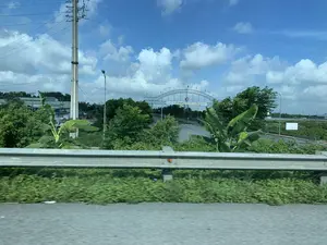 Bản ghi văn bản trên cổng chào lớn là 'KHU CÔNG NGHIỆP ĐỒNG VĂN II MỞ RỘNG', bên dưới có dòng chữ nhỏ hơn không thể đọc rõ. Ngoài ra còn có biển báo giới hạn tốc độ '30'. Bức ảnh chụp cảnh quan từ một phương tiện đang di chuyển trên đường, hướng về lối vào một khu công nghiệp. Địa điểm là khu vực gần đường cao tốc, tại cổng vào Khu công nghiệp Đồng Văn II Mở Rộng. Hoạt động chính là di chuyển trên đường; ở phía xa có một máy xúc màu vàng cho thấy có thể đang diễn ra hoạt động xây dựng. Các vật thể nổi bật bao gồm cổng chào lớn bằng kết cấu thép có treo cờ phướn, lan can hộ lan bằng kim loại ở tiền cảnh, cây cối xanh tươi ven đường (có cả cây chuối), các cột điện cao thế, đèn đường, và các tòa nhà kiểu nhà xưởng ở phía xa, dưới bầu trời xanh có mây trắng.。（日本語訳）。大きなウェルカムゲートの上の文字は「KHU CÔNG NGHIỆP ĐỒNG VĂN II MỞ RỘNG」（ドンバンII拡張工業団地）であり、その下には小さくてはっきりと読めない文字の列がある。また、「30」という速度制限の標識もある。この写真は、道路を移動中の車両から工業団地の入口に向かって撮影された風景である。場所は高速道路近くのエリアで、ドンバンII拡張工業団地の入口ゲートである。主な活動は道路上の移動である。遠くには黄色いショベルカーがあり、建設活動が行われている可能性を示している。