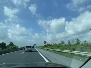 Bức ảnh chụp lại khung cảnh giao thông trên một tuyến đường cao tốc ở Việt Nam, nhìn từ trong một chiếc ô tô. Nhiều xe ô tô đang lưu thông trên đường, trong đó có một chiếc xe màu bạc ở phía trước. Bầu trời trong xanh và có nhiều mây trắng. Hai bên đường là hàng rào bảo vệ và cây cối xanh tươi. Có các biển báo giao thông bên lề đường. Một biển báo hình tròn viền đỏ ghi số '100', cho biết tốc độ tối đa cho phép là 100 km/h. Ngay bên dưới là biển báo hình tam giác cảnh báo có công trình đang thi công. Ở phía xa hơn, có một biển báo lớn màu xanh lá cây chỉ dẫn lối ra: 'LỐI RA PHỦ LÝ 3km', và chỉ hướng đi 'TP. PHỦ LÝ', 'QUỐC LỘ 1A'.。（日本語訳）。ベトナムのある高速道路の交通風景を、一台の車内から撮影した写真です。多くの自動車が道路を走行しており、その中には前方に銀色の車が1台います。空は青く澄んでおり、白い雲が多くあります。道路の両側にはガードレールと青々とした木々があります。路肩には交通標識があります。赤い縁の円形標識には「100」と書かれており、最高許容速度が時速100kmであることを示しています。そのすぐ下には、工事中を警告する三角形の標識があります。さらに遠くには、「フーリー出口 3km」と出口を案内する大きな緑色の標識があり、「フーリー市」、「国道1A号線」への方向を示しています。
