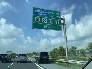 Đây là hình ảnh chụp từ trong một chiếc ô tô đang di chuyển trên đường cao tốc ở Việt Nam vào một ngày nắng đẹp, trời xanh có mây. Phía trước là một biển báo giao thông lớn màu xanh lá cây, chỉ dẫn đến trạm dừng nghỉ. Nội dung trên biển báo ghi rõ: "TRẠM DỪNG NGHỈ DUY TIÊN", "REST AREA", "Km 227+000", và khoảng cách còn lại là "1,5 km". Biển báo còn có các biểu tượng cho biết trạm dừng nghỉ có các dịch vụ: trạm xăng, nhà hàng ăn uống, nhà vệ sinh và khu vực sửa chữa xe. Trên đường có một số xe ô tô khác đang lưu thông. Hai bên đường cao tốc là hàng cây xanh và lan can bảo vệ. Ở phía xa bên phải, có một biển báo màu xanh dương ghi "ĐỊA PHẬN TP. PHỦ LÝ ZONE", cho biết khu vực này thuộc địa phận thành phố Phủ Lý.。（日本語訳）。これは、晴れた美しい日、青空に雲が浮かぶベトナムの高速道路を走行中の車内から撮影された画像です。前方には、サービスエリアへの案内を示す大きな緑色の交通標識があります。標識には、「TRẠM DỪNG NGHỈ DUY TIÊN」（ズイティエン・サービスエリア）、「REST AREA」、「Km 227+000」、そして残りの距離が「1.5 km」と明記されています。標識には、サービスエリアにガソリンスタンド、レストラン、トイレ、車両修理エリアといったサービスがあることを示すピクトグラムもあります。道路上には、他にも数台の自動車が走行しています。高速道路の両側には、緑の木々の列とガードレールがあります。遠く右側には、「ĐỊA PHẬN TP. PHỦ LÝ ZONE」（フーリー市 区域）と書かれた青い標識があり、この地域がフーリー市の管轄区域であることを示しています。