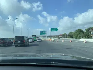 Bức ảnh chụp lại khung cảnh giao thông trên một tuyến đường cao tốc ở Việt Nam, nhìn từ trong một chiếc ô tô. Nhiều phương tiện như ô tô con, xe khách và xe van đang lưu thông trên đường. Có các biển báo giao thông cung cấp thông tin cho tài xế. Một biển báo lớn màu xanh lam ghi 'TRẠM DỪNG NGHỈ CT. CẦU GIẼ NINH BÌNH', cho biết trạm dừng nghỉ cách đó 750m. Một biển báo khác màu xanh lá cây chỉ lối ra 'TP. PHỦ LÝ' và 'QUỐC LỘ 1A'. Phía dưới biển báo trạm dừng nghỉ có biển hiệu của 'TỔNG CÔNG TY ĐẦU TƯ PHÁT TRIỂN ĐƯỜNG CAO TỐC VIỆT NAM'. Gần lối ra, có một biển báo tạm thời màu vàng ghi chữ 'CHẬM' và các rào chắn bê tông được đặt trên đường, cho thấy có thể đang có công trình thi công. Phía xa có một cây cầu vượt. Bầu trời trong xanh và có nhiều mây.。（日本語訳）。ベトナムのある高速道路の交通風景を車内から撮影した写真です。乗用車、バス、バンなど多くの車両が道路を通行しています。ドライバーに情報を提供する交通標識があります。「TRẠM DỪNG NGHỈ CT. CẦU GIẼ NINH BÌNH」と書かれた大きな青い標識があり、サービスエリアが750m先にあることを示しています。別の緑色の標識は「TP. PHỦ LÝ」と「QUỐC LỘ 1A」への出口を示しています。サービスエリアの標識の下には、「TỔNG CÔNG TY ĐẦU TƯ PHÁT TRIỂN ĐƯỜNG CAO TỐC VIỆT NAM」（ベトナム高速道路開発投資総公社）の看板があります。出口付近には、「CHẬM」（徐行）と書かれた黄色い仮設標識と、道路に設置されたコンクリート製の防護柵があり、工事が行われている可能性を示しています。遠くには陸橋があります。空は青く澄んでおり、多くの雲が浮かんでいます。